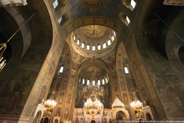 Interior of Alexander Nevsky Cathedral looking upward toward painted domes, arched openings, icons, and hanging chandeliers.