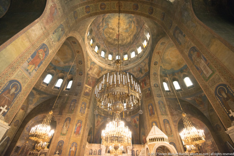 Upward view of Alexander Nevsky Cathedral’s painted dome and large central chandelier framed by mural-covered arches.