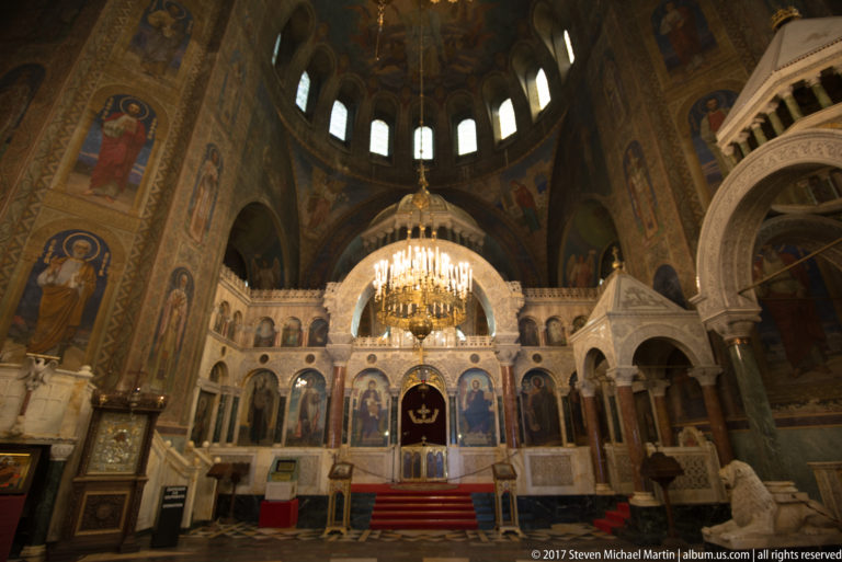 Alexander Nevsky Cathedral sanctuary with pale iconostasis, large chandelier, painted saints, and red-carpeted steps.