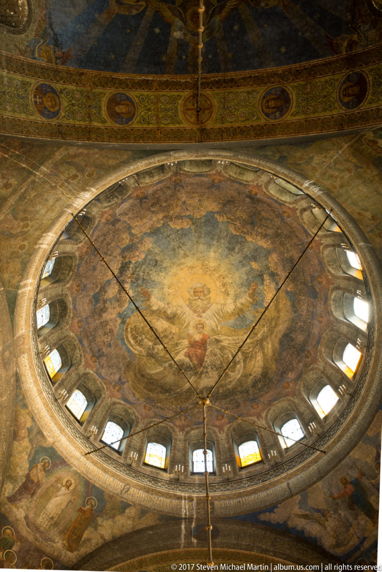 Upward view of a painted cathedral dome encircled by windows and decorative bands inside Alexander Nevsky Cathedral.