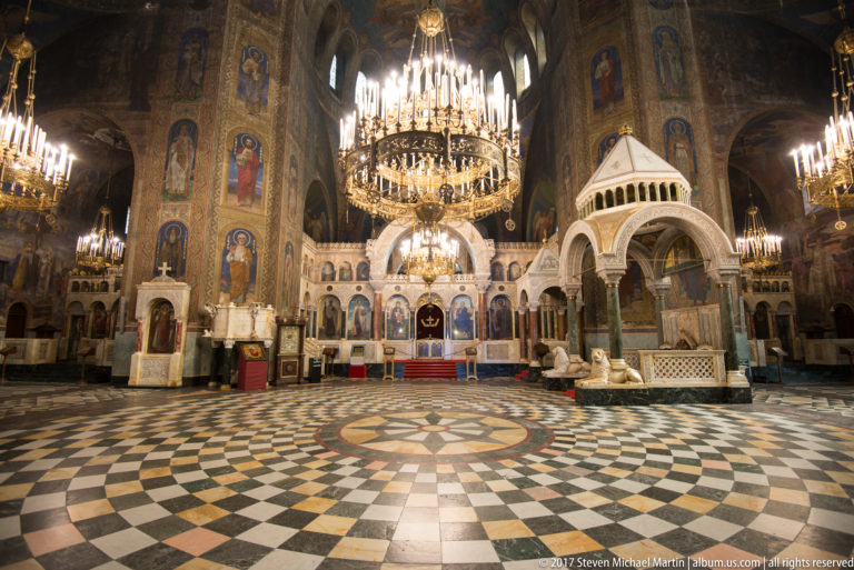 Wide sanctuary interior with checkered marble floor, chandeliers, iconostasis, and painted walls at Alexander Nevsky Cathedral.