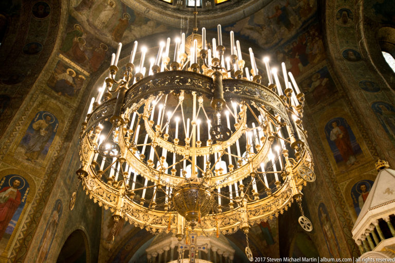 Large multi-tiered gilded chandelier with many candle-shaped lights hanging before painted walls in Alexander Nevsky Cathedral.