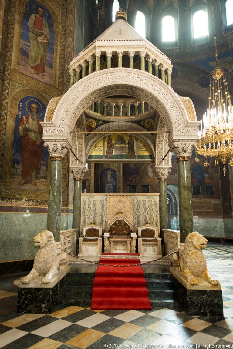 White carved marble canopy with green columns, red-carpeted steps, and sculptural details inside Alexander Nevsky Cathedral.