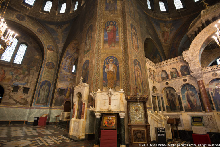 Tall pier painted with saints beside carved white sanctuary architecture and devotional stands in Alexander Nevsky Cathedral.