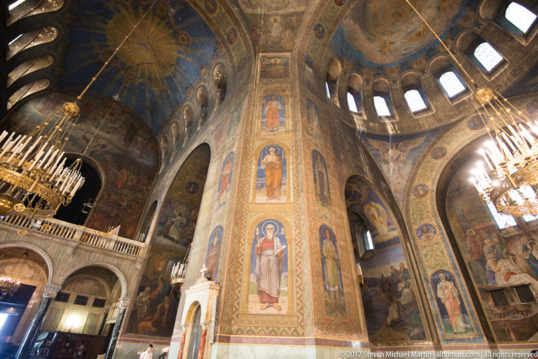 Upward view of a tall pier painted with saints beneath domes, arched windows, and chandeliers in Alexander Nevsky Cathedral.