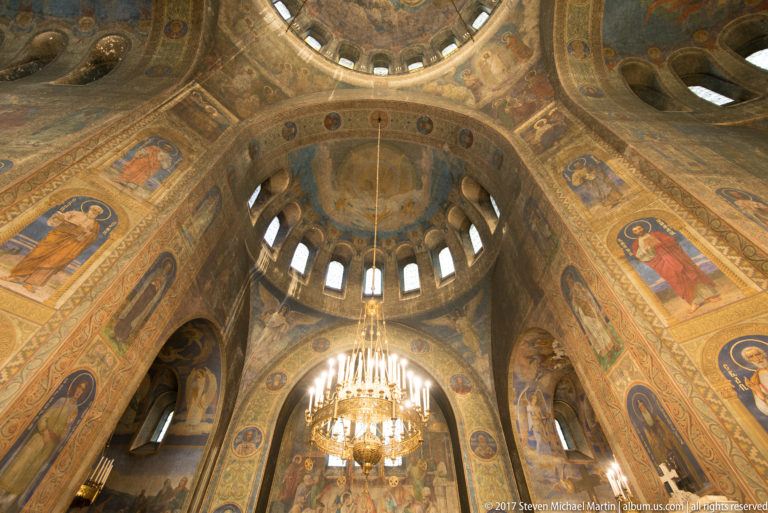Painted central dome with bright windows, surrounding saint murals, and a chandelier inside Alexander Nevsky Cathedral.