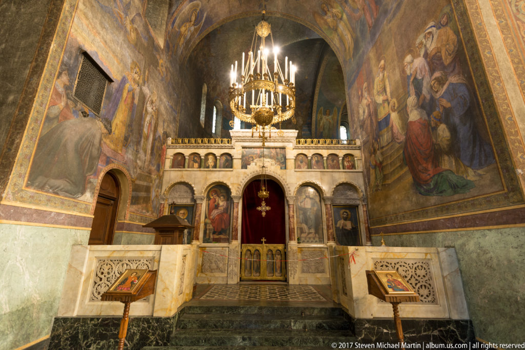 Smaller liturgical area with carved stone rails, icon screen, chandelier, and large wall paintings in Alexander Nevsky Cathedral.