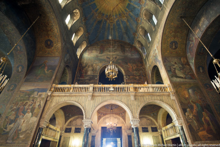 Rear gallery on pale arches beneath mural-covered vaults and a blue-toned dome inside Alexander Nevsky Cathedral.
