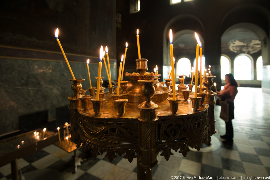 Ornate metal candle stand holding many lit taper candles inside a dim Orthodox cathedral interior.