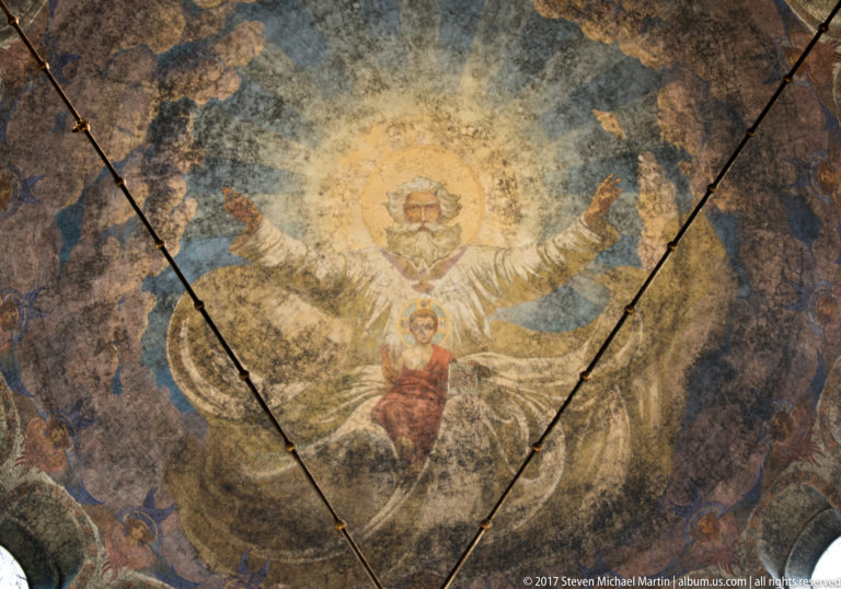 Weathered Orthodox ceiling fresco with a haloed Christ figure, smaller sacred figure, blue field, and visible surface cracks.