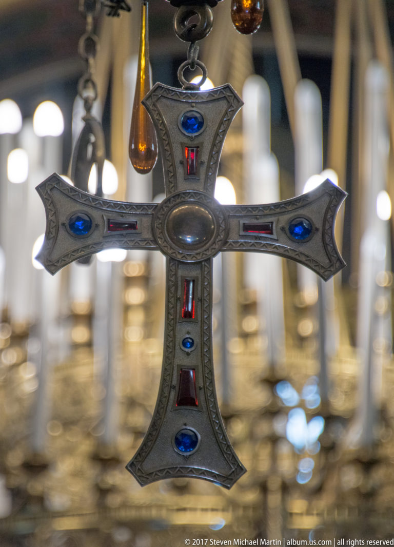 Decorative metal Orthodox cross with blue and red inset stones hanging before blurred church lamps and candlesticks.