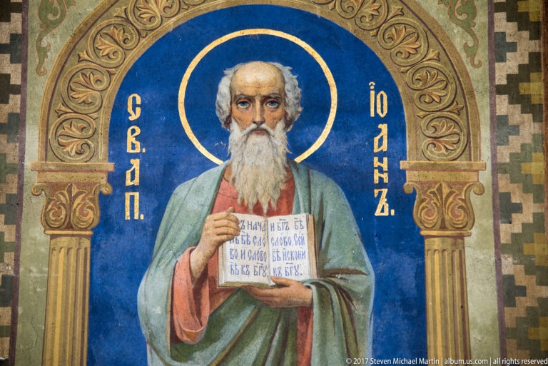 Mosaic icon of an elderly bearded saint with gold halo holding an inscribed book against a blue background.