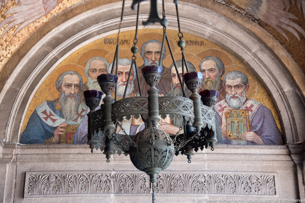 Semicircular mosaic of bearded clerical figures above an arch, partly obscured by a hanging metal chandelier.