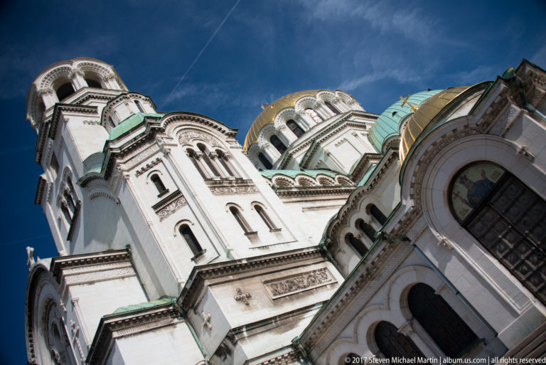 Low-angle view of Alexander Nevsky Cathedral with white façades, arched windows, green roofs, towers, and gilded dome.
