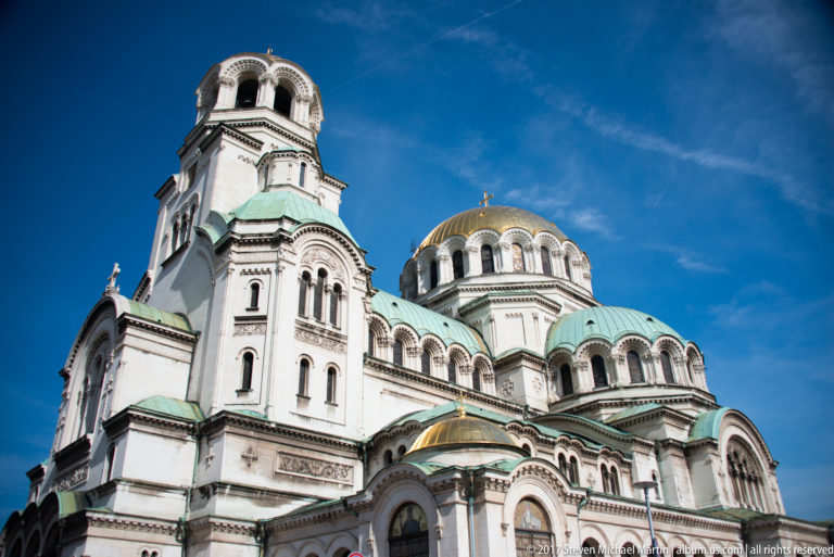 Alexander Nevsky Cathedral façade with tall bell tower, green roofs, arched windows, and gilded central dome under blue sky.