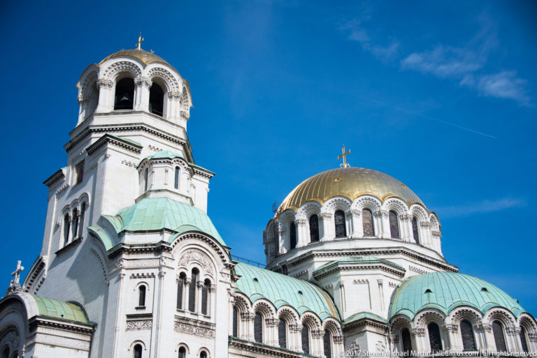 Upper section of Alexander Nevsky Cathedral with open-arched bell tower, gilded dome, and green-patinated roofs.