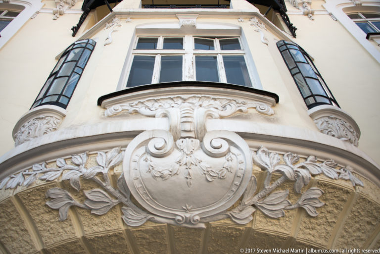 Upward view of an ornate pale façade with sculpted cartouche, floral stucco, curved bay, and narrow windows.