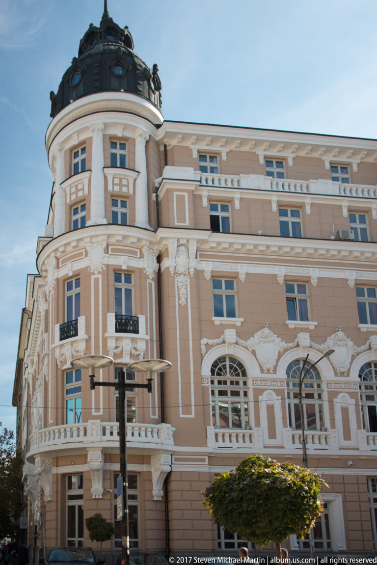 Pink-and-cream historic corner building with rounded façade, white ornament, tall windows, balconies, trees, and streetlight.