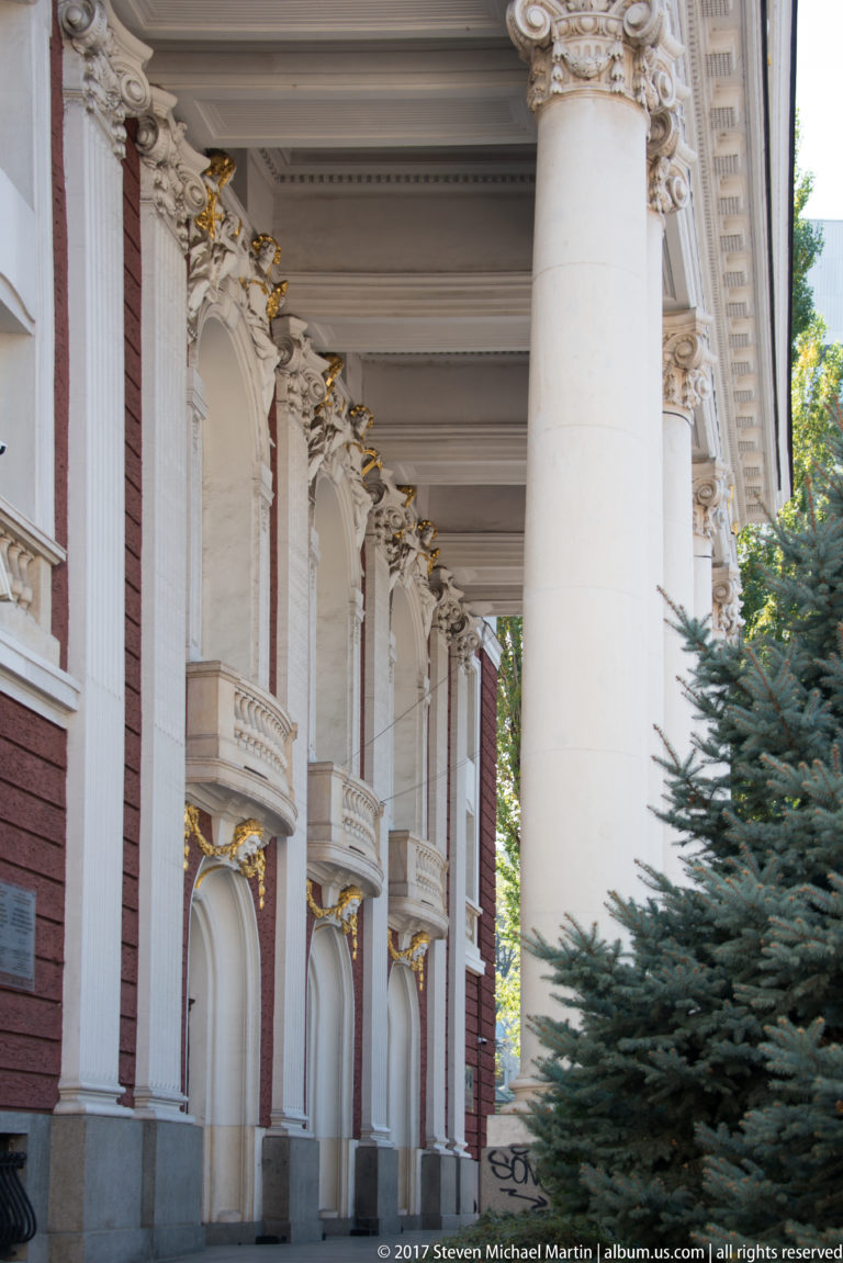 Side colonnade of Ivan Vazov National Theatre with tall white columns, red walls, arches, gilded details, and evergreen trees.