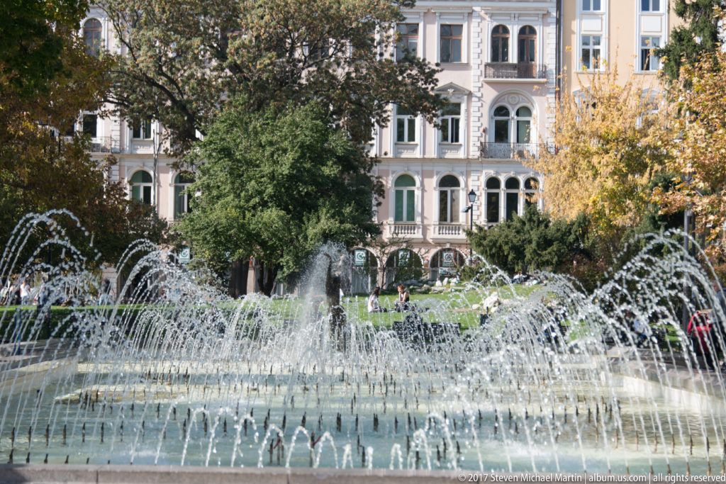 Curving fountain jets in Sofia’s City Garden with trees and a historic multi-story building in the background.
