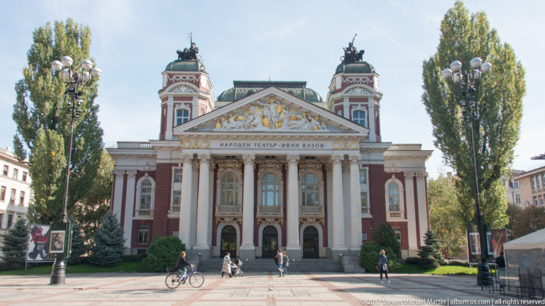 Front view of Ivan Vazov National Theatre with columned portico, sculpted pediment, twin towers, trees, pedestrians, and cyclist.