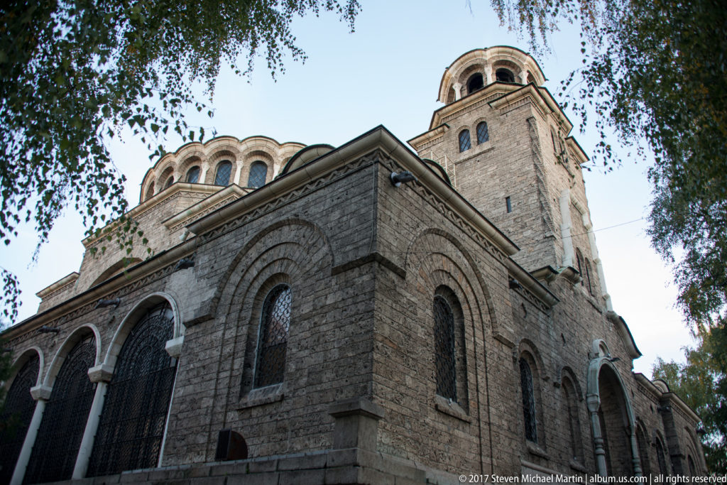 Low-angle exterior of Saint Nedelya Church with gray stone walls, arched windows, domed roofline, bell tower, and trees.