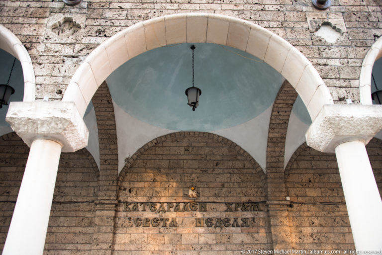 Cyrillic wall inscription on exposed brick beneath an arched portico with white columns and a hanging lamp.