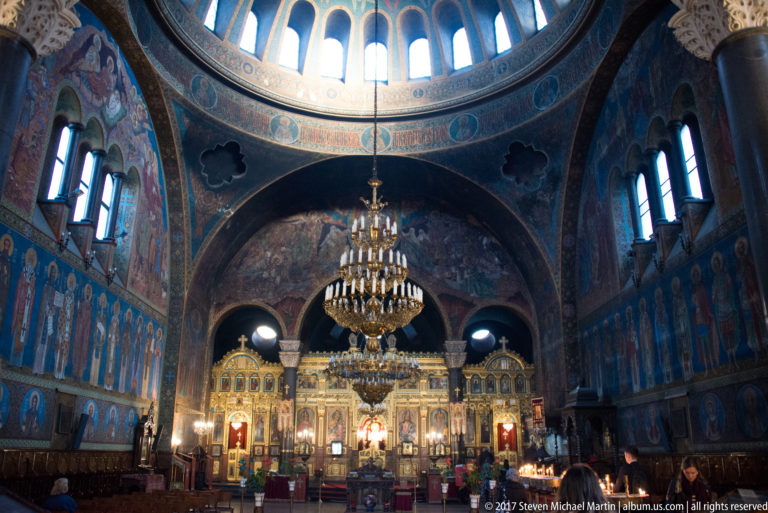 Interior of Saint Nedelya Church with gilded iconostasis, chandeliers, blue frescoed walls, arches, and high clerestory windows.