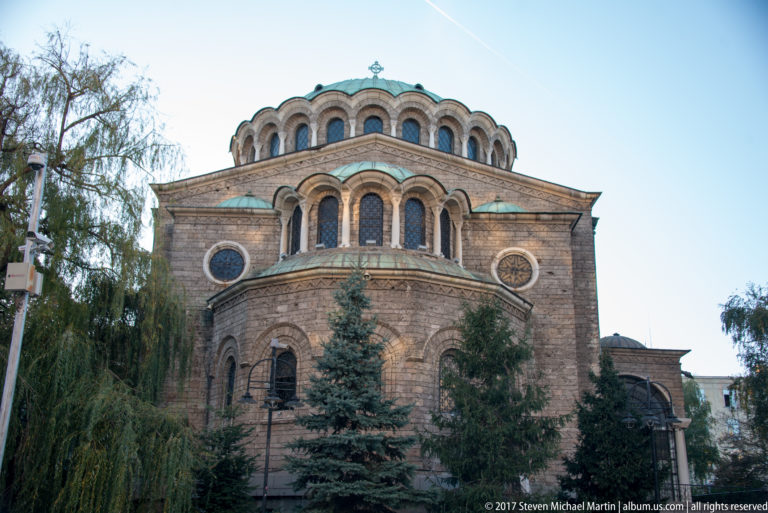 Rear exterior of Saint Nedelya Church with layered masonry, arched upper windows, round windows, green roof, and surrounding trees.