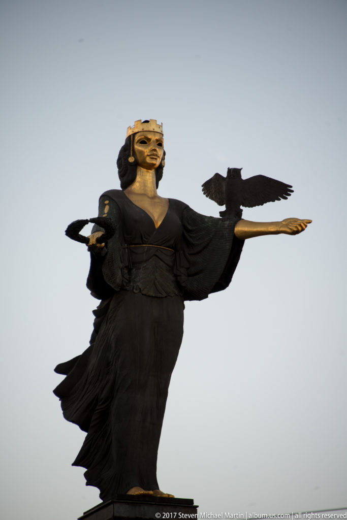 Upper figure of the Statue of Sofia with dark drapery, gilded face, outstretched arms, and bird against the sky.