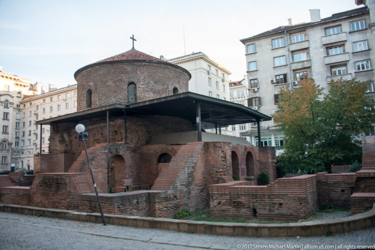 Brick Rotunda of St. George with conical roof and exposed archaeological masonry, surrounded by modern buildings in central Sofia.
