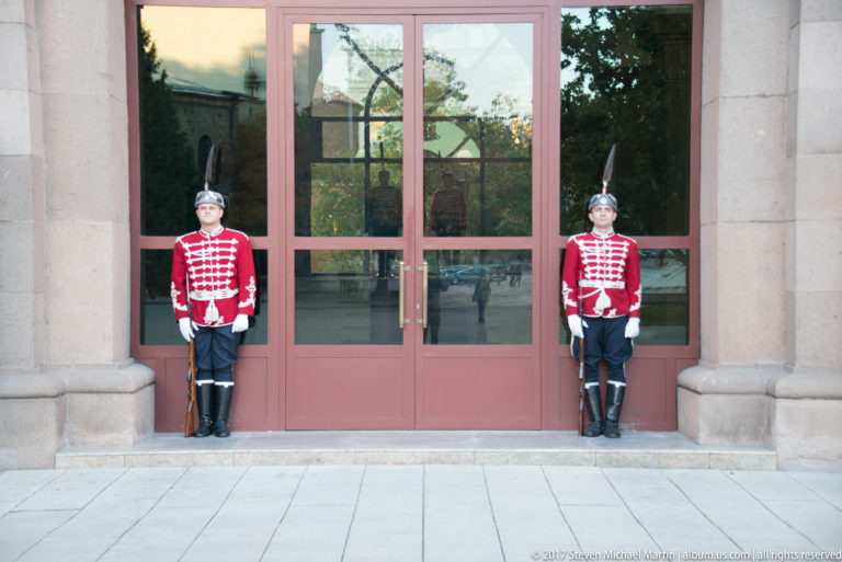 Two Bulgarian ceremonial guards in red uniforms stand with rifles beside a glazed stone entrance.
