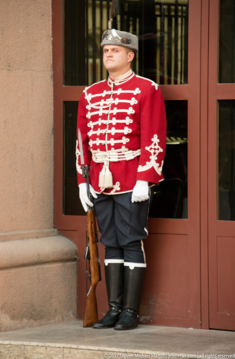 Single Bulgarian ceremonial guard in red braided tunic, helmet, white gloves, black boots, and rifle at a doorway.