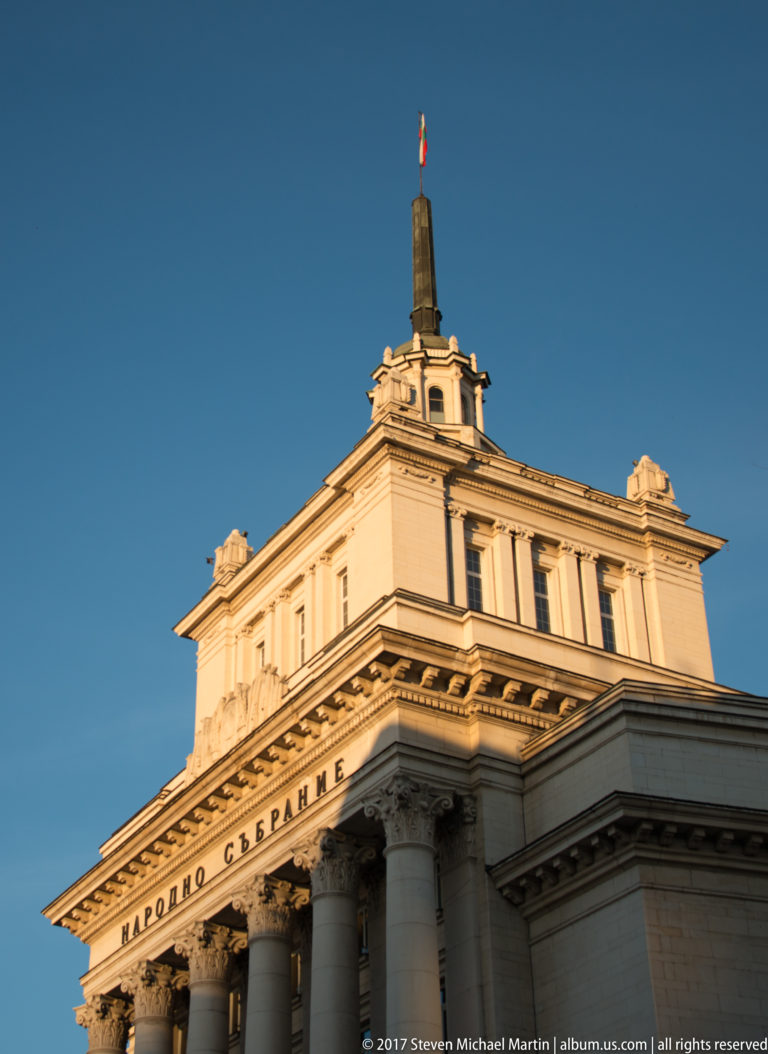 Upper façade of Bulgaria’s National Assembly with classical columns, Bulgarian inscription, rooftop flag, and clear blue evening sky.