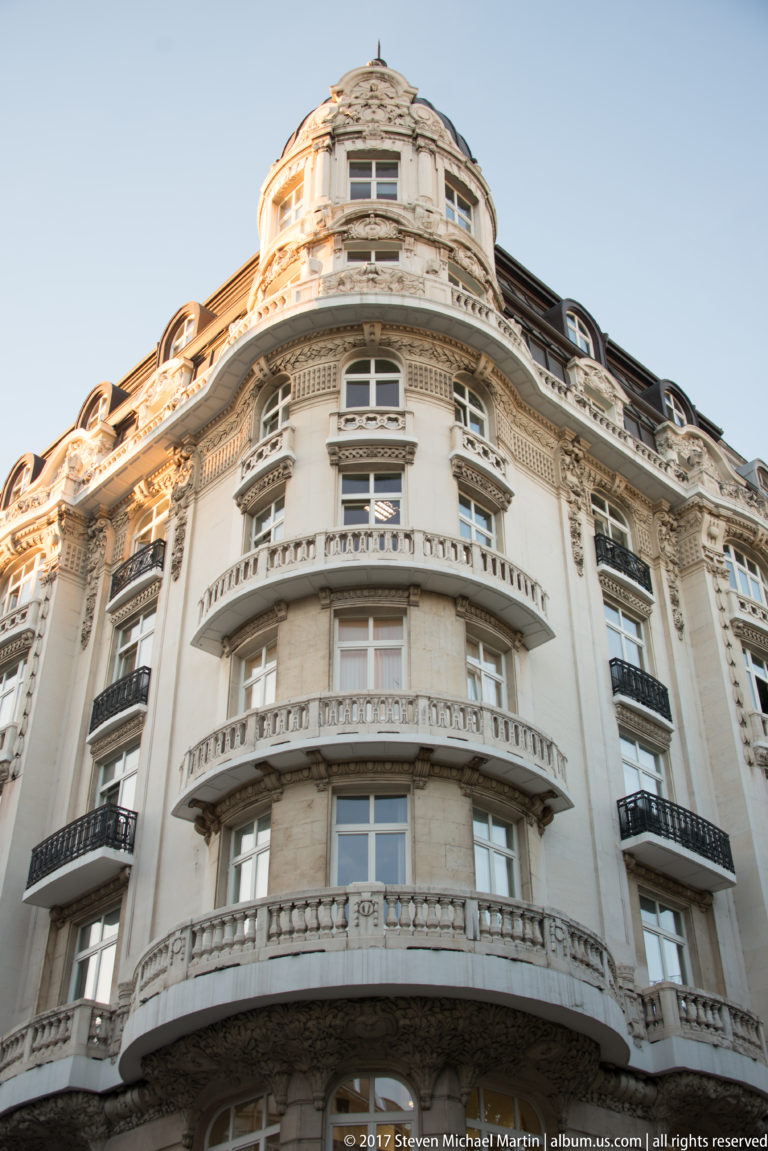 Low-angle view of an ornate curved corner building with stacked balconies, tall windows, and decorative pale stone façade.