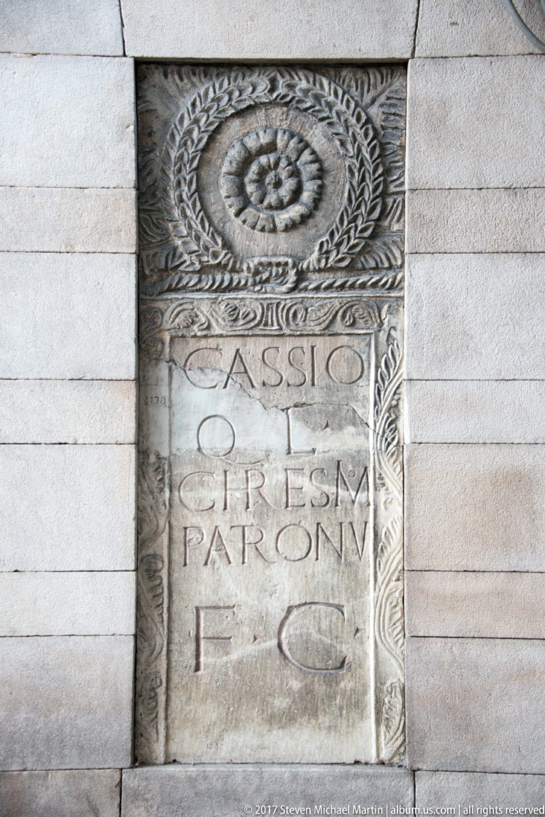 Carved Roman stone fragment with circular ornament, Latin inscription, and decorative border embedded in a masonry wall.