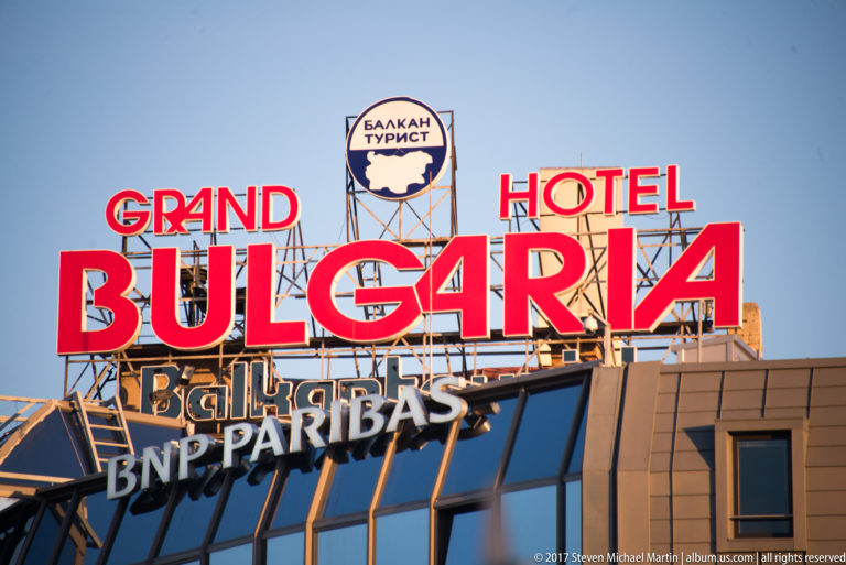 Large red rooftop sign reading Grand Hotel Bulgaria above smaller commercial signs against a clear sky.