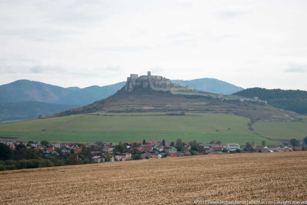 Distant hilltop castle above fields and a village, with wooded mountain ridges in the background.