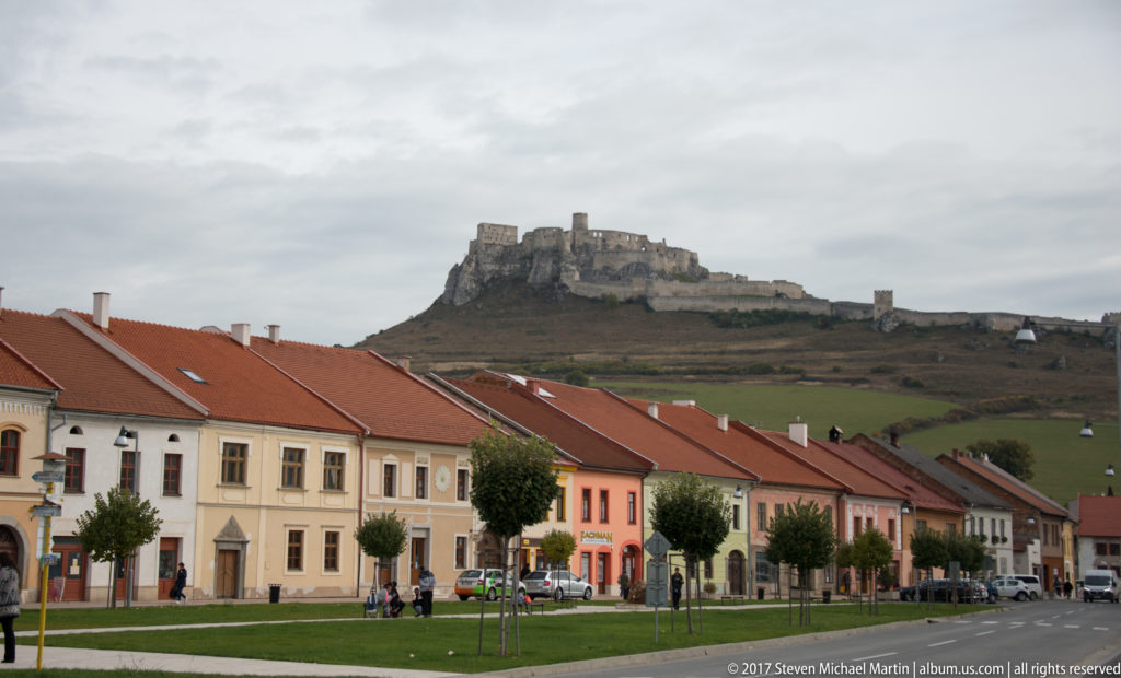 Pastel town houses and red roofs below Spiš Castle, which rises on a hill in the background.
