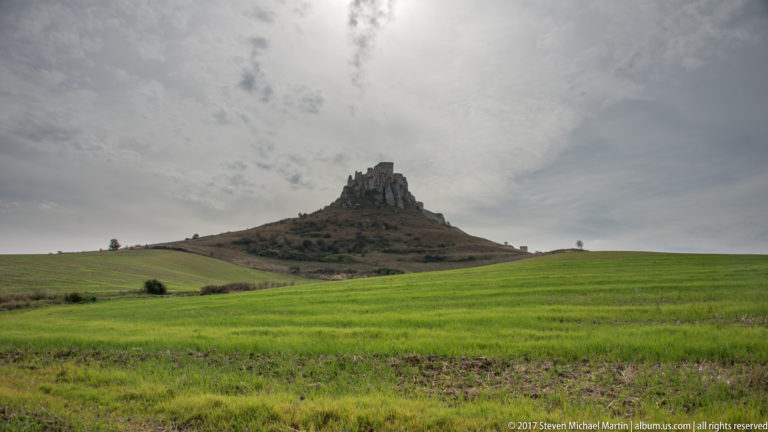 Distant stone ruin on a rocky hill above broad green fields beneath a cloudy sky.