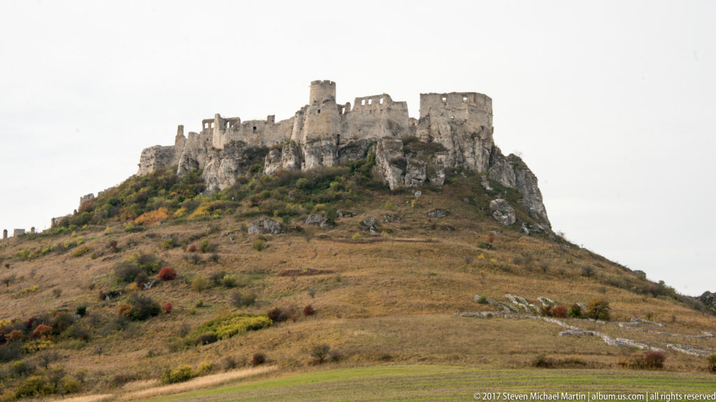Spiš Castle ruins extending across a rocky hilltop above dry grass and autumn shrubs.