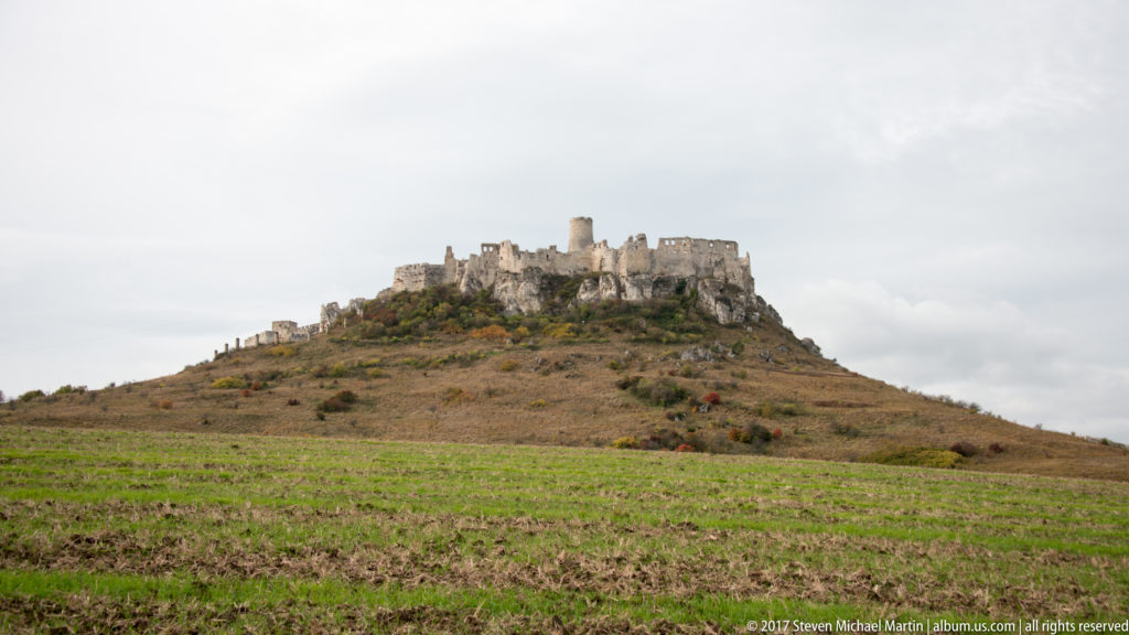Wide view of Spiš Castle on a tawny hill beyond green cultivated rows under an overcast sky.
