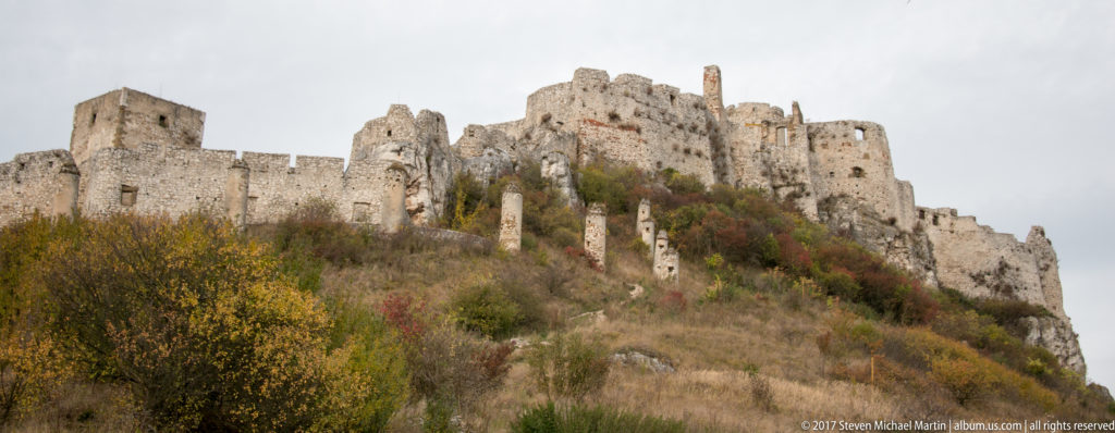 Upward view of Spiš Castle's massive ruined walls and towers above a steep shrub-covered slope.