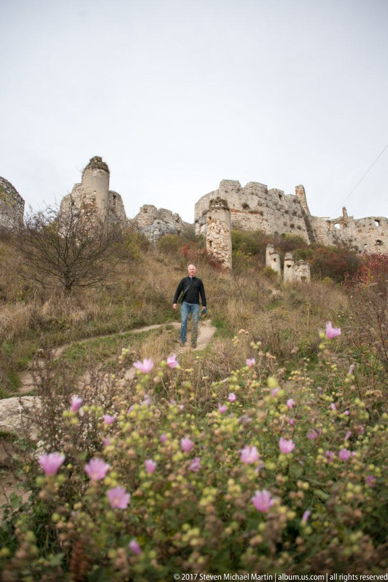 Person walking through pink wildflowers and rough grass below low ruined stone walls on a castle hillside.