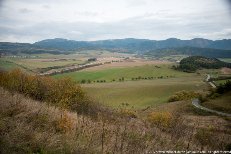 Elevated valley view over patchwork fields, roads, and forested ridges, with dry hillside vegetation in front.