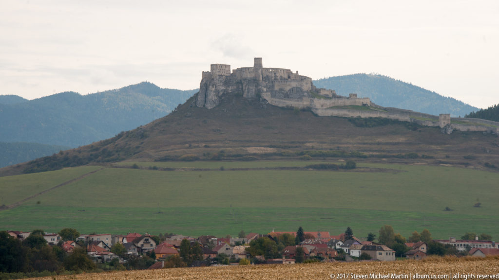Hilltop castle above green fields and red-roofed village houses, backed by dark mountain slopes.