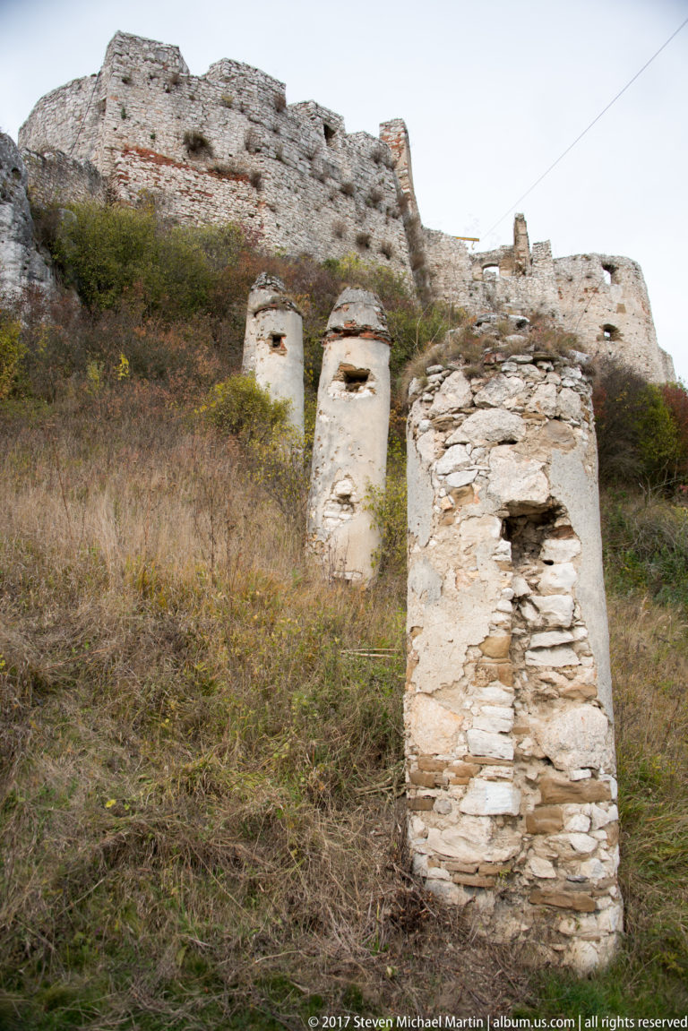 Three weathered freestanding masonry piers with small recesses standing in grass below ruined castle walls.