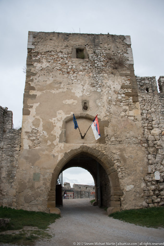 Round-arched gateway through a tall weathered castle wall, with two flags above and a courtyard beyond.