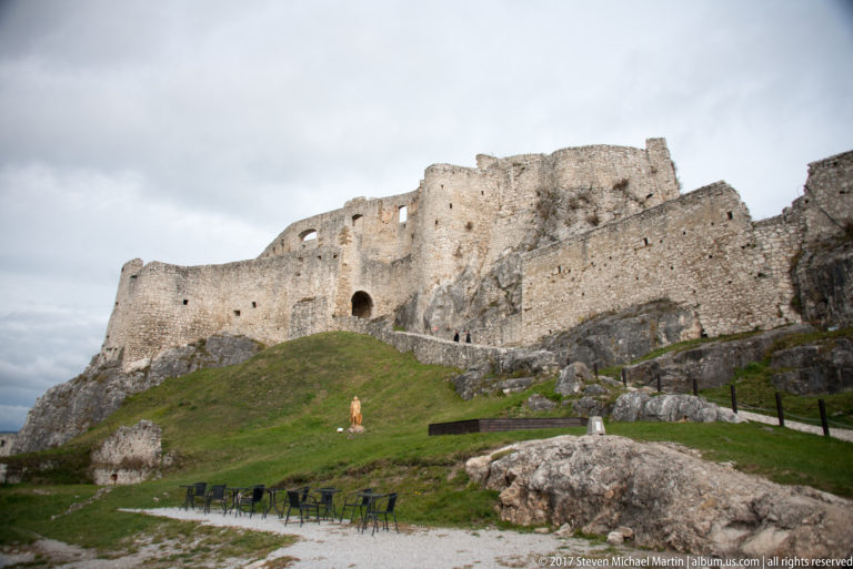 Massive ruined stone walls beside a grassy lower court and ascending path inside Spiš Castle.