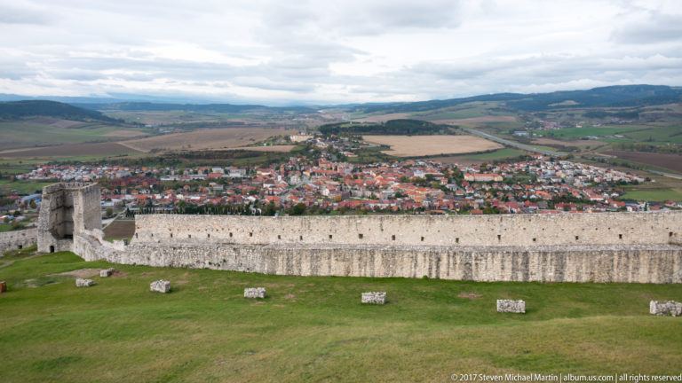 Long castle wall above a grassy enclosure, with a red-roofed town, fields, and hills extending across the valley beyond.